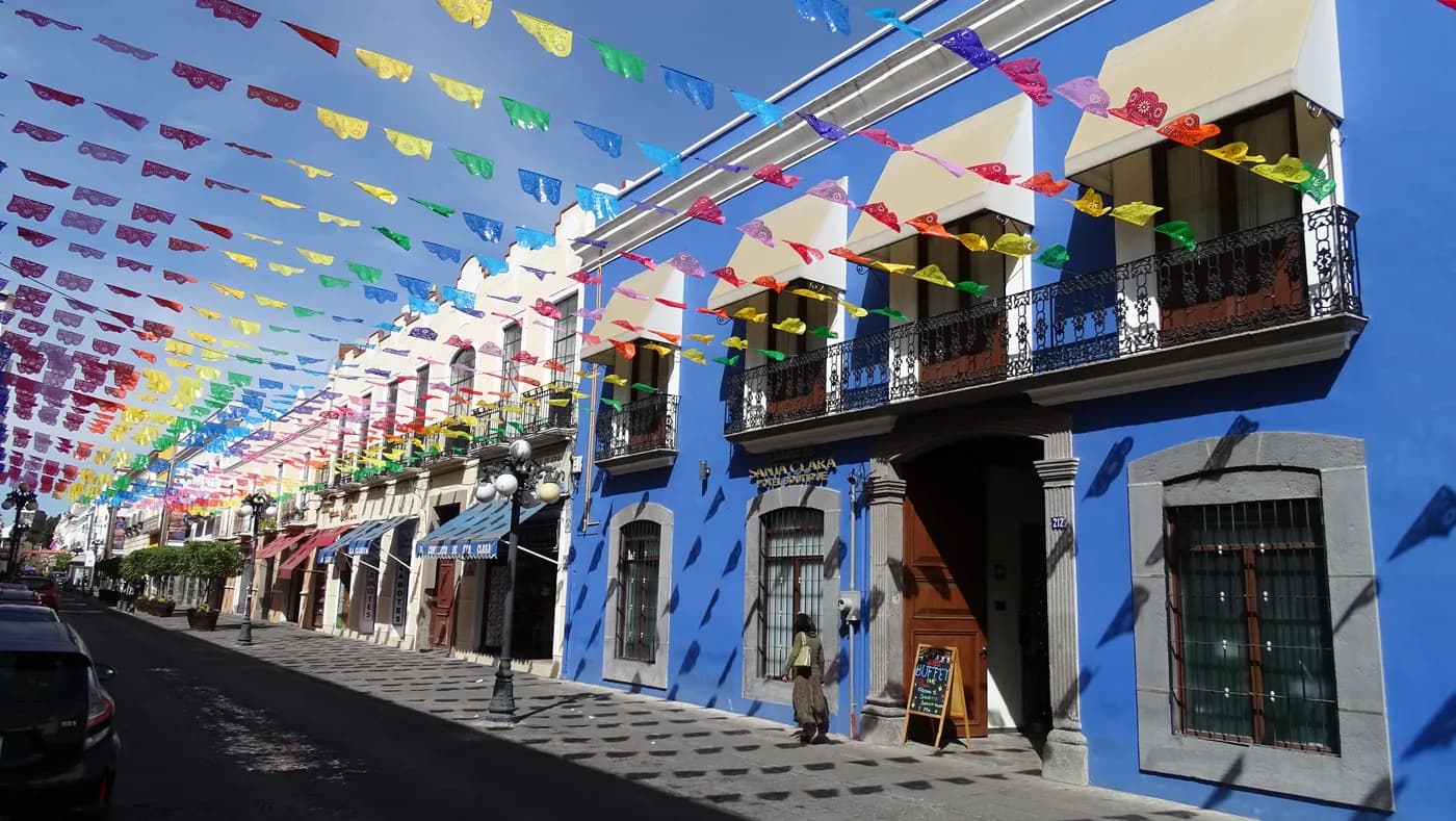 Colorful papel picado hangs above blue colonial buildings on a sunlit street in Puebla, Mexico